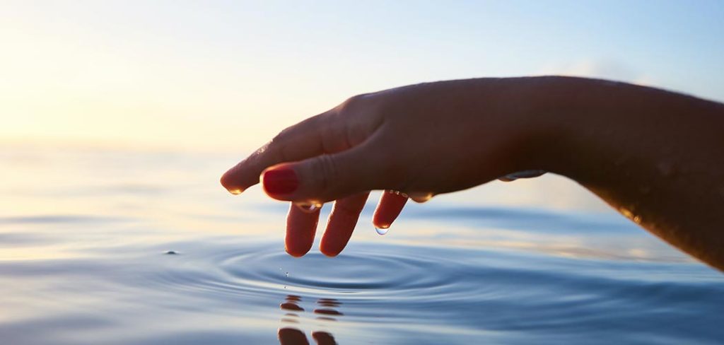 A dripping bare hand and forearm hovers over a body of water that blends at the horizon with the sky