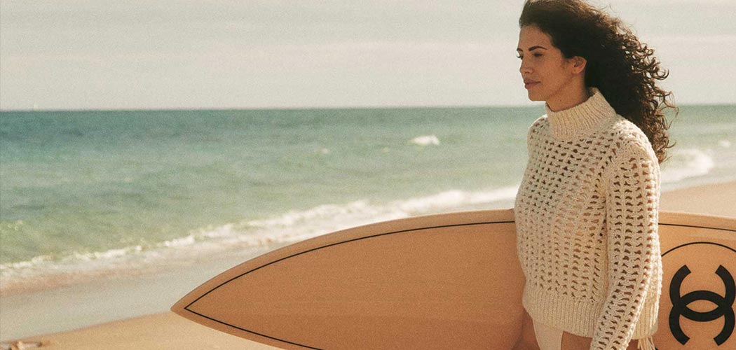 Hope Dworaczyk in a white sweater on a beach holding a Chanel surfboard
