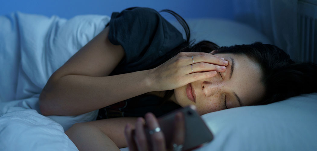 An image of a woman laying down on her side in bed, rubbing her eyes and holding her cellphone