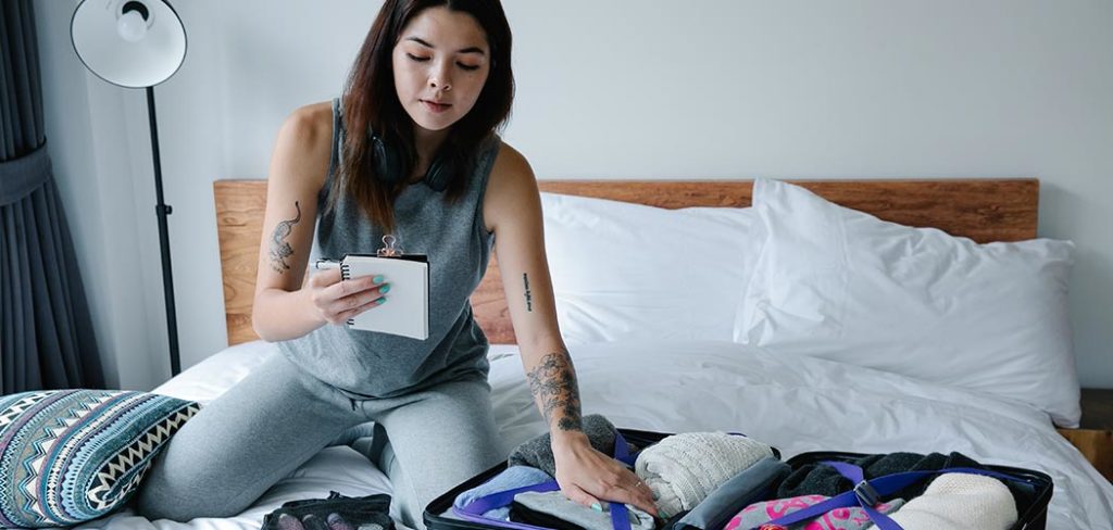 A brunette woman with tattoos on her arms checks a list while packing her suitcase on a bed.