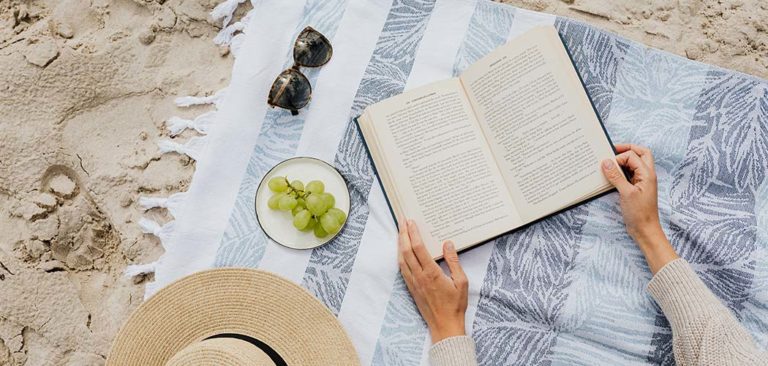 An image of an open book laying on a beach towel surrounded by sunglasses, a bowl of grapes, and a sun hat.
