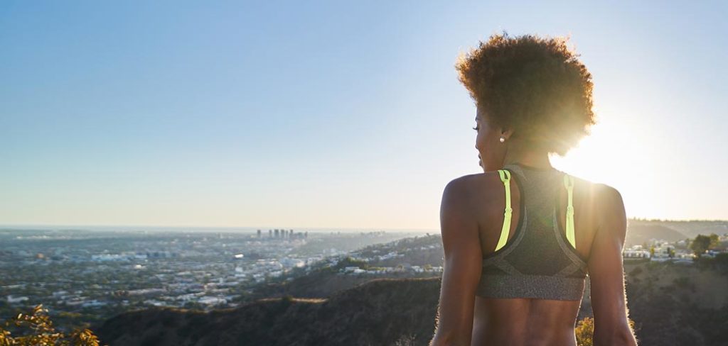 An African American woman rests and looks out on a city vista while wearing workout clothes.