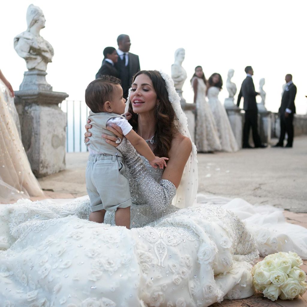Hope Dworaczyk in a white wedding dress kneeling down on the ground and holds a child