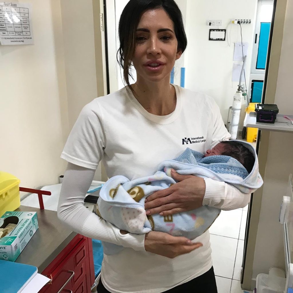 Hope Dworaczyk wears a white shirt with an International Medical Corps logo as she holds an infant at a refugee camp in Jordan