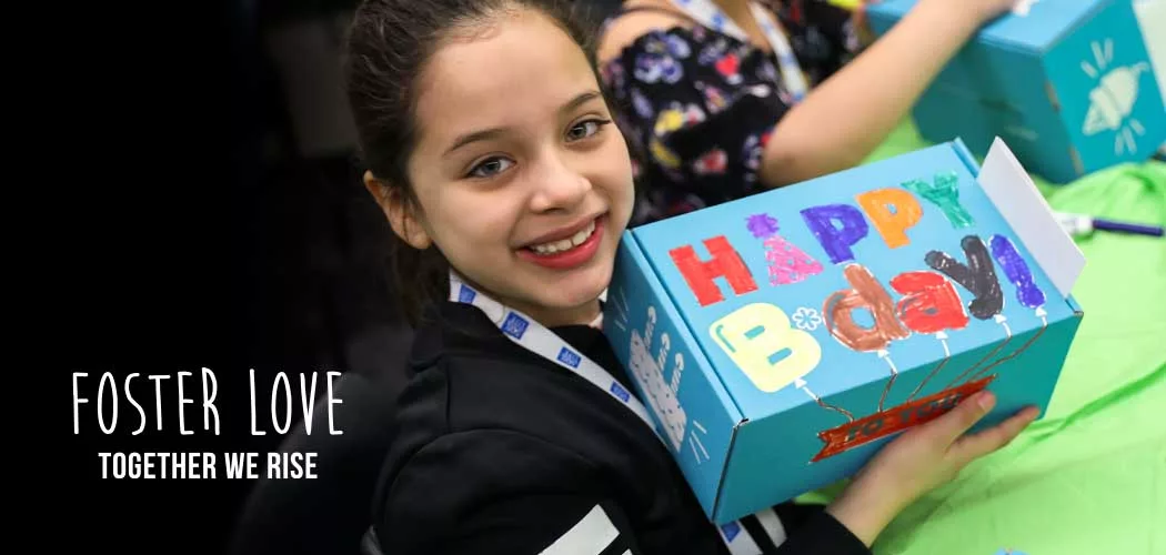 A child smiling while holding a blue shoebox with “happy bday” on the front. The Together We Rise logo is in the bottom right corner.