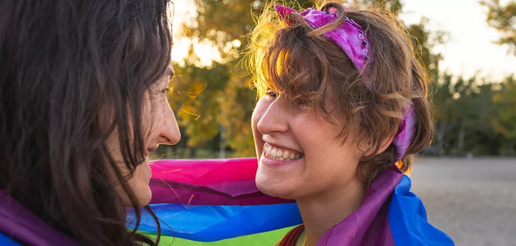 Two people smiling at each other while they are wrapped in the pride flag.