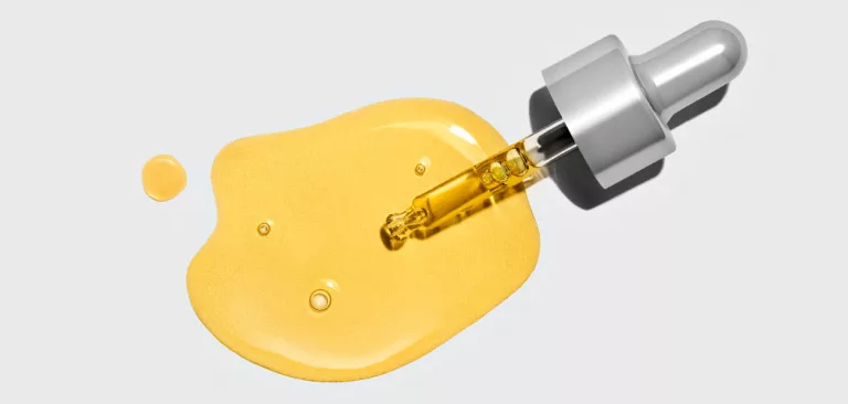 A makeup dropper full of yellow product dripping out over a white surface.